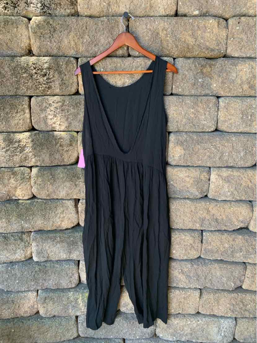 Black sleeveless jumpsuit with deep scoop neck and loose, gathered wide legs hanging on wooden hanger against stone wall.