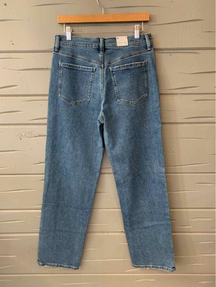 Blue denim high-waisted jeans with straight legs and back pockets hanging on wooden hanger against beige wall.