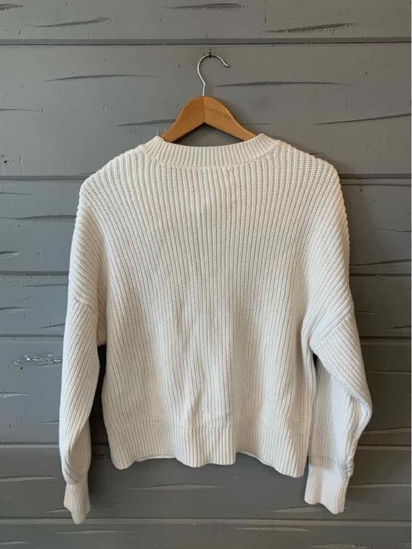 White ribbed knit sweater with long sleeves, crew neckline, and relaxed fit hanging on wooden hanger against gray wooden.