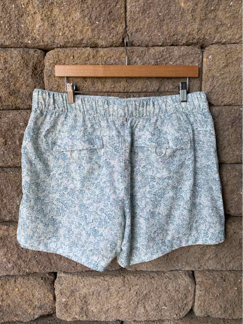 Light blue floral patterned shorts with elastic waistband hanging on wooden hanger against stone wall.