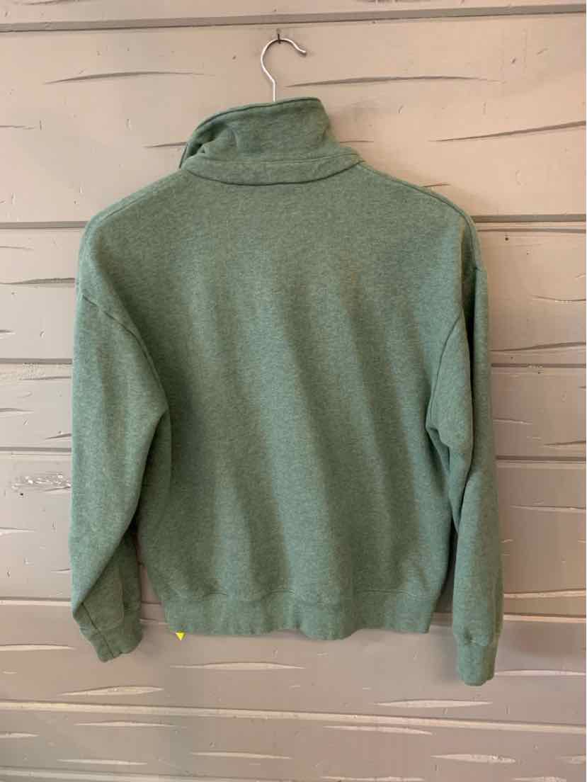 Green long-sleeve pullover with high collar and ribbed cuffs and hem hanging on white wooden wall.