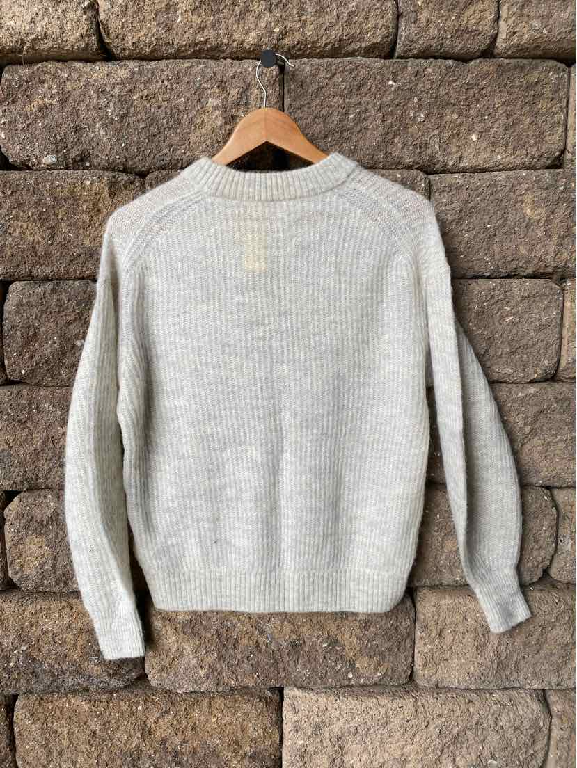 Light gray knit sweater with long sleeves and ribbed cuffs hanging on wooden hanger against stone wall.