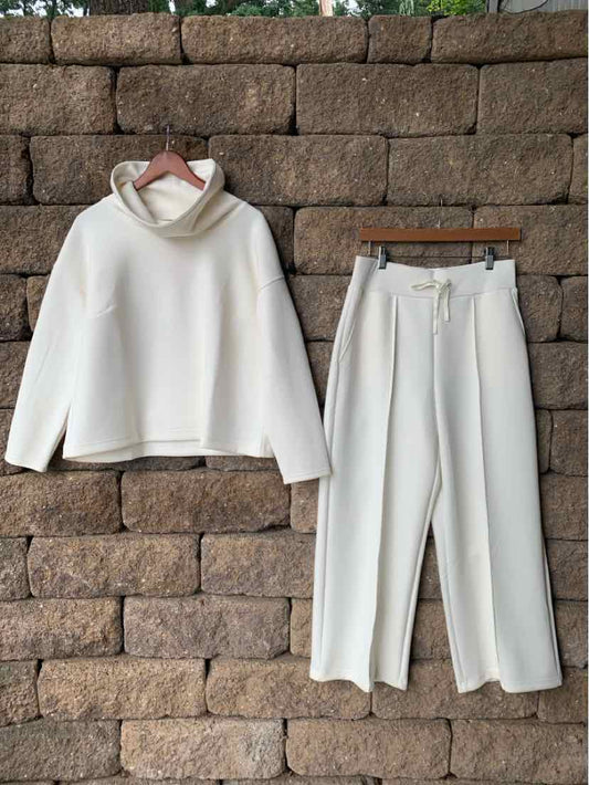Cream-colored two-piece loungewear set with a loose turtleneck sweatshirt and matching drawstring pants hanging against a.