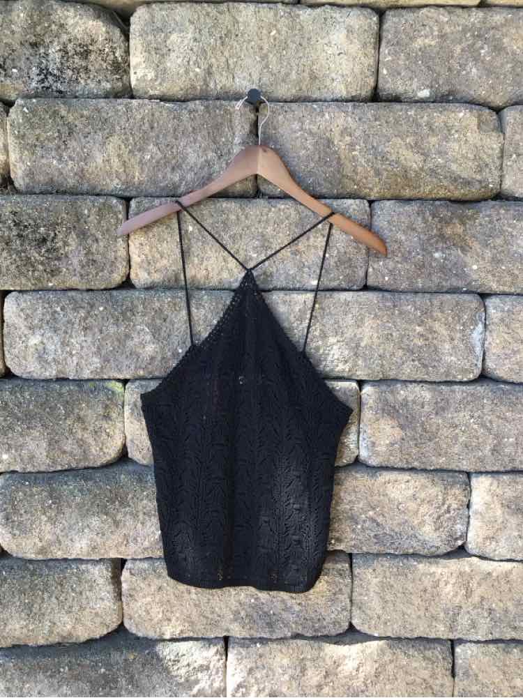 Black crochet tank top with thin crisscross straps hanging on wooden hanger against stone wall.