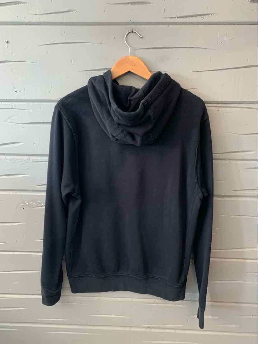 Black long-sleeve hoodie with a front pocket and relaxed fit on wooden hanger against gray wooden wall.