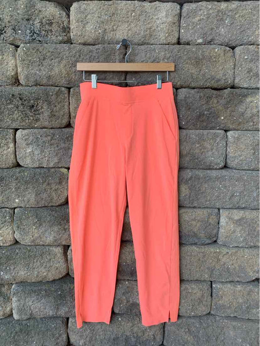 Coral-orange tapered pants with high waist and ankle zippers hanging on wooden hanger against stone wall.