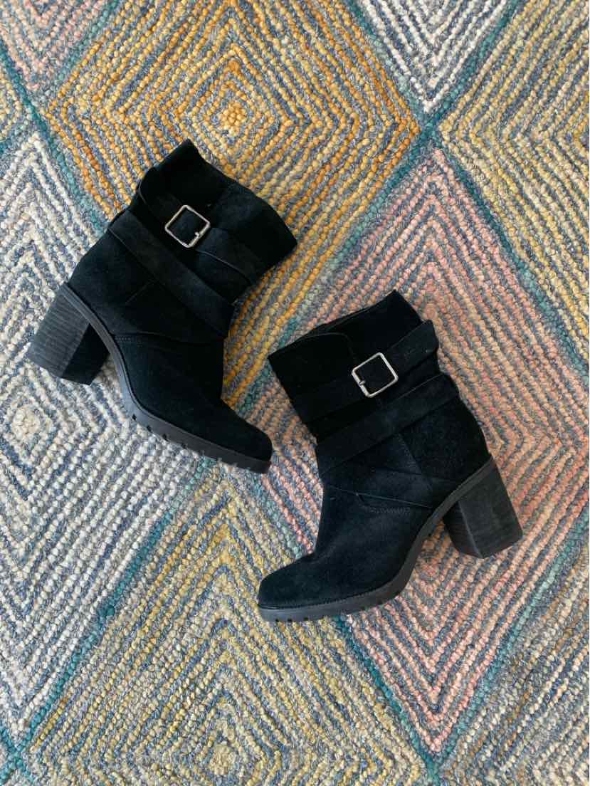 Black suede ankle boots with block heels and side buckle detail on textured multicolor rug.