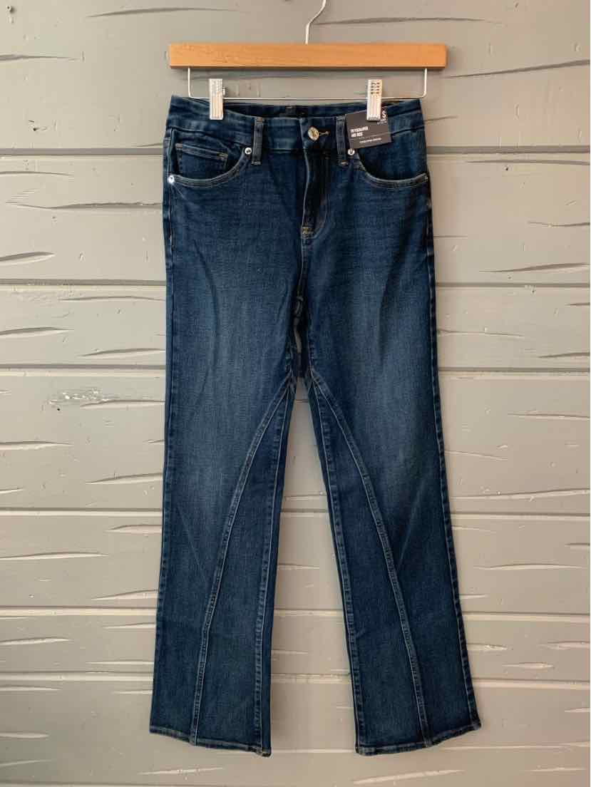 Dark blue slim-fit denim jeans with front pockets and button closure hanging on wooden hanger against gray wall.