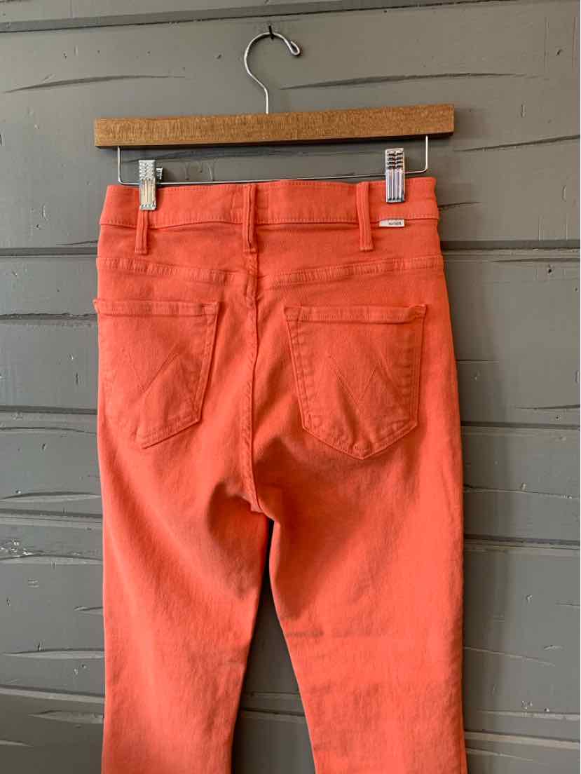 Orange high-waist jeans with back pockets and belt loops on wooden hanger against gray wall.