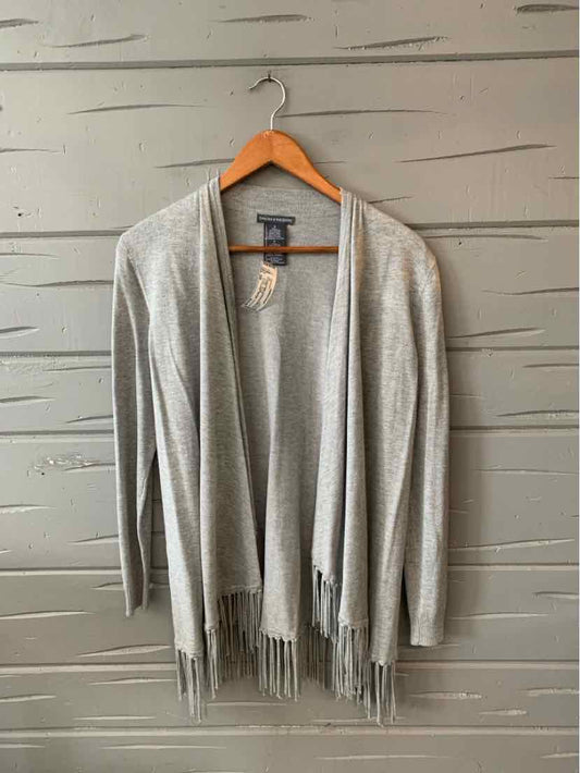 Light gray open-front cardigan with long sleeves and fringe hem hanging on wooden hanger against gray wood panel wall.