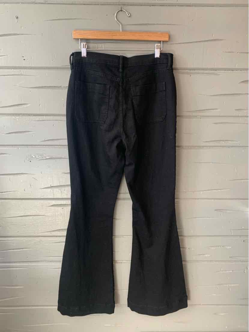 Black denim pants with slight flare, back view, size 10, hanging on wooden hanger against light wooden wall.