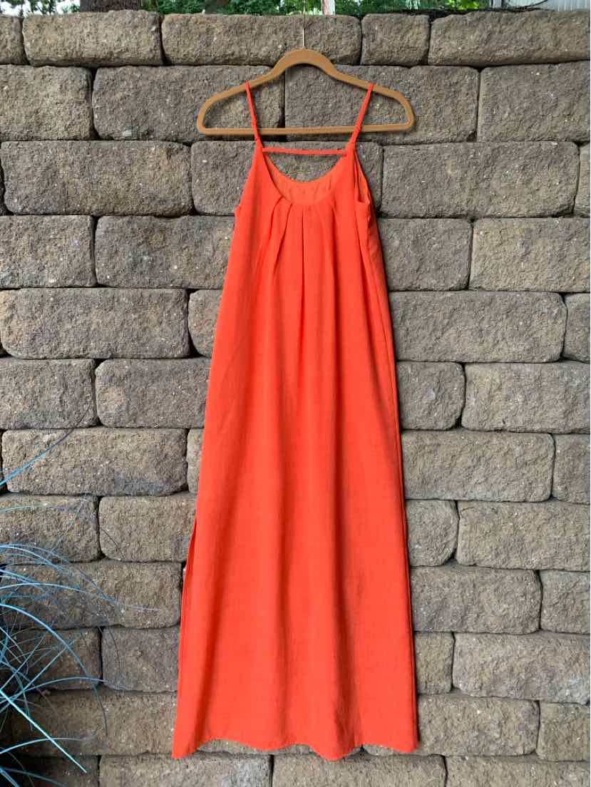 Bright orange sleeveless dress with thin straps and a loose, flowy fit hanging on a wooden hanger against a stone wall.