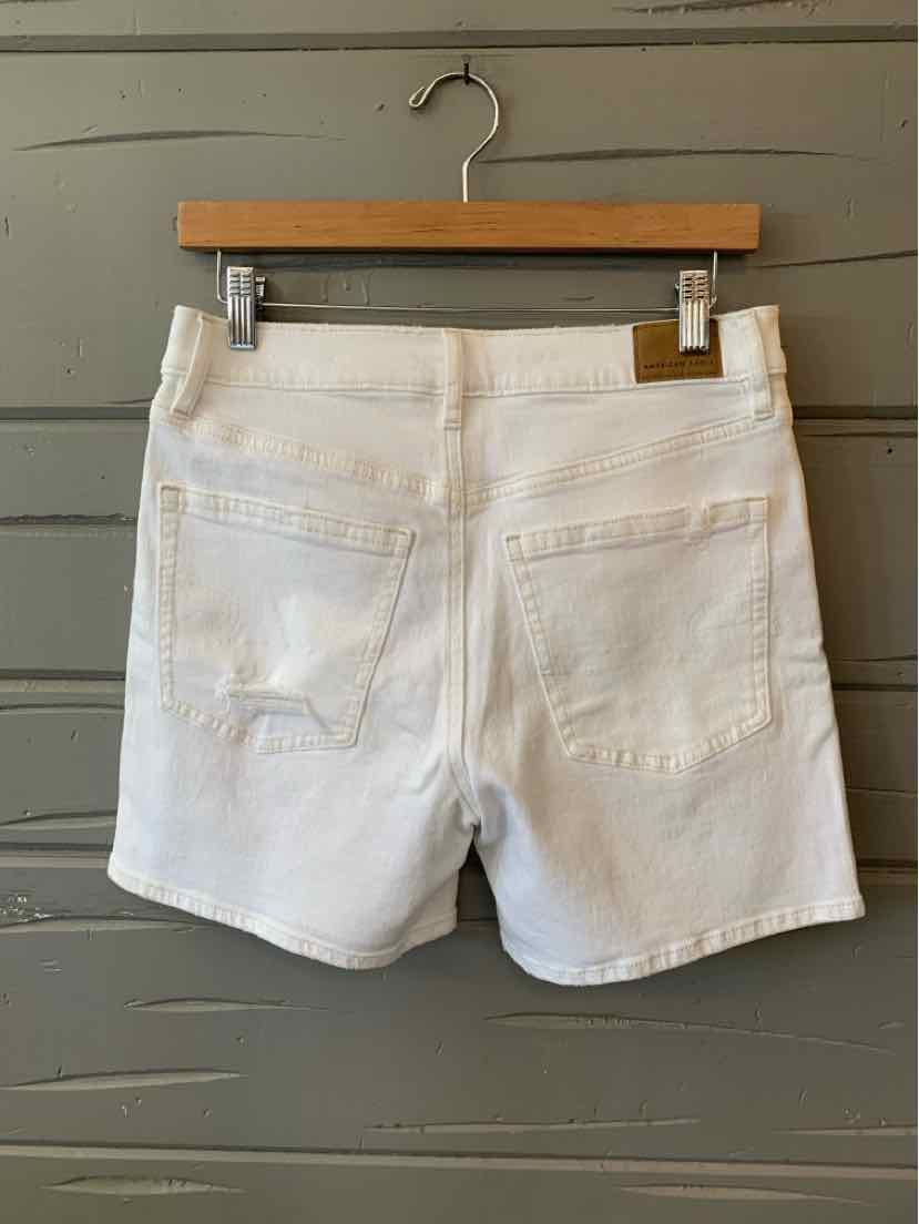 White denim shorts with back pockets and belt loops hanging on wooden hanger against gray wood background.