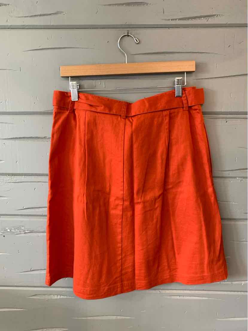 Bright orange knee-length skirt with a fitted waistband and side zipper closure hanging on a wooden hanger against a gray.