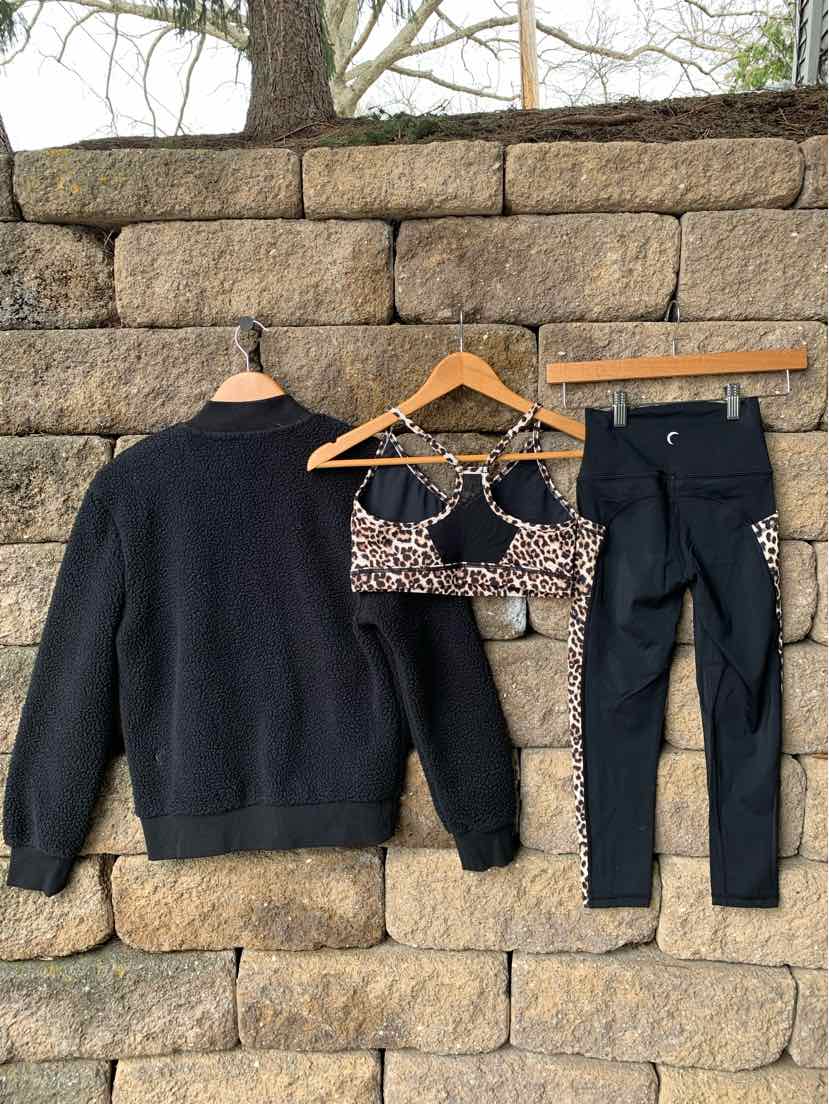 Black sherpa jacket, leopard print sports bra with crisscross straps, and black leggings with zipper pockets hanging on a.