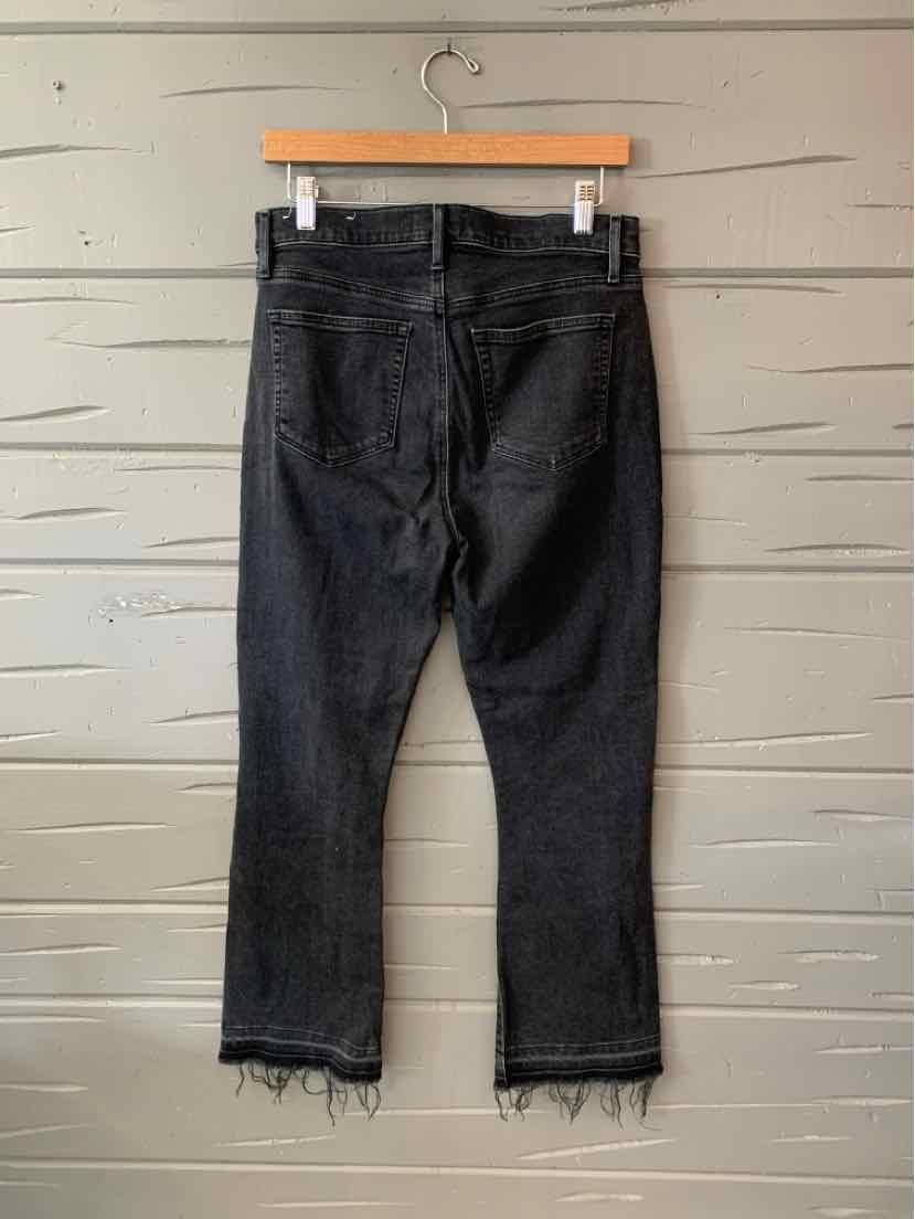 Black denim cropped jeans with raw frayed hems and back pockets hanging on wooden hanger against gray wall.