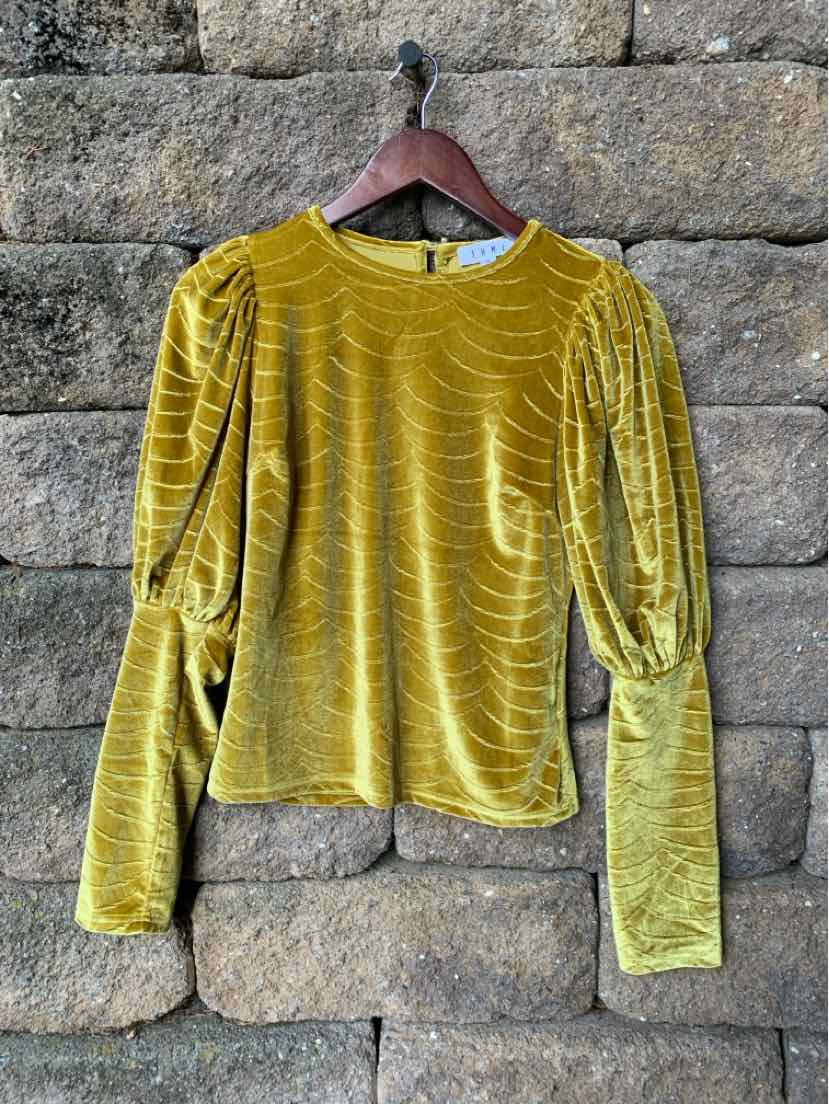 Mustard yellow velvet long-sleeve top with puffed shoulders and fitted cuffs on wooden hanger against stone wall.