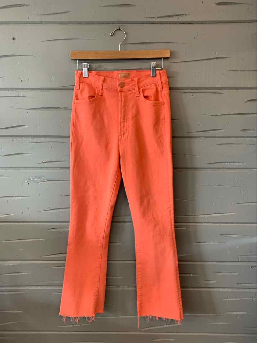 Orange high-waist jeans with raw hem and front pockets hanging on wooden hanger against gray wooden background.