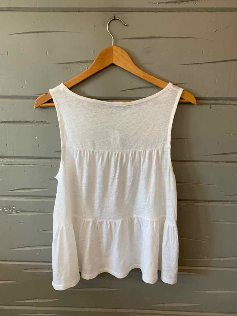 White sleeveless tiered blouse with scoop neckline on wooden hanger against gray wooden wall.