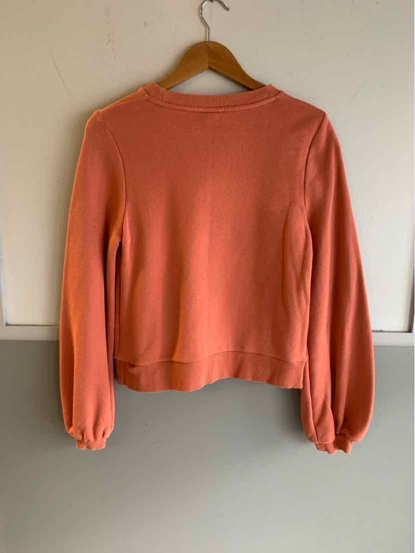 Coral orange long-sleeve pullover with ribbed cuffs and hem on wooden hanger against white wall.