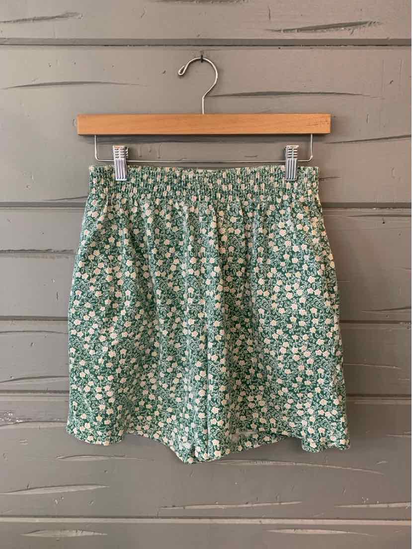 Green floral high-waist shorts with elastic waistband and loose fit hanging on wooden hanger.