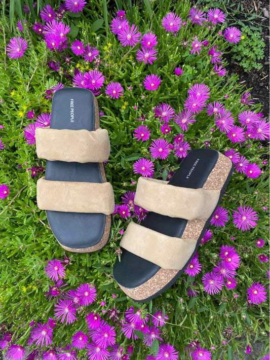 Tan double-strap sandals with black footbed and cushioned sole on grass with purple flowers.