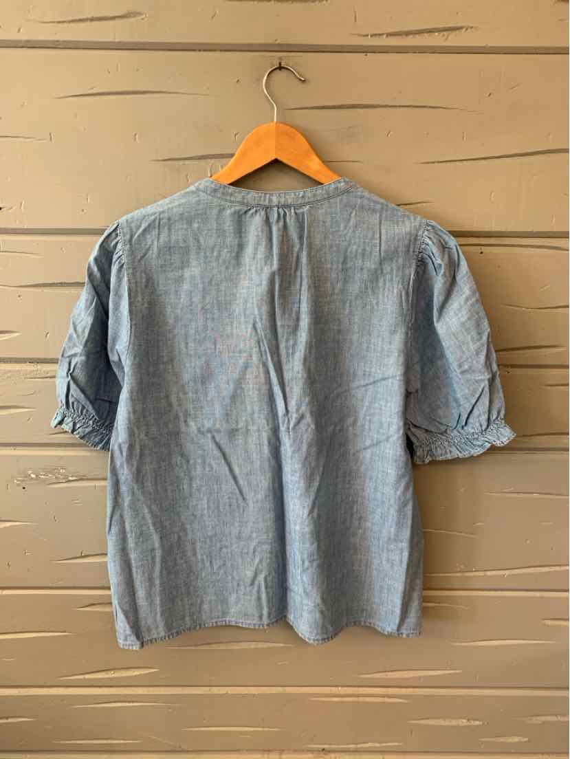 Light blue chambray blouse with three-quarter sleeves and a relaxed fit hanging on wooden hanger.