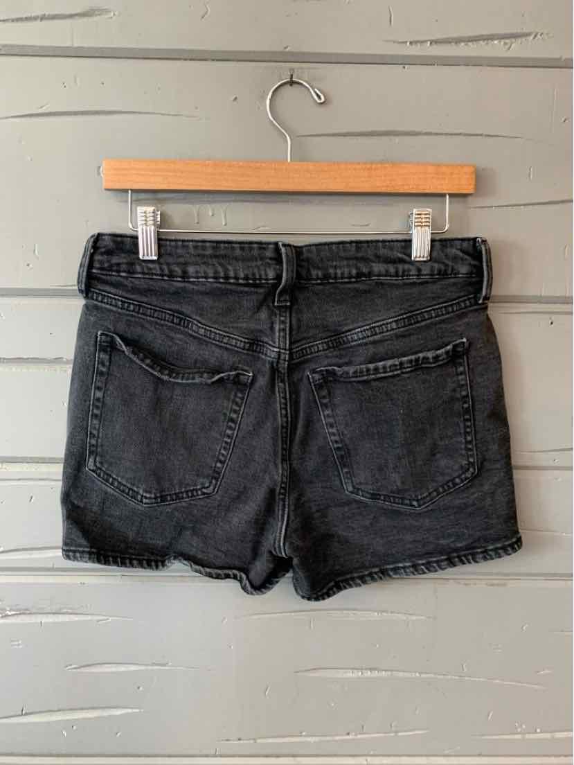 Black denim shorts with distressed hem and two back pockets hanging on wooden hanger against gray wood wall.