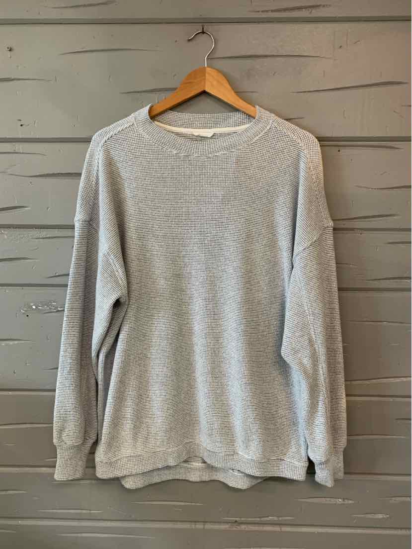 Light gray crewneck sweatshirt with long sleeves and ribbed cuffs hanging on wooden hanger against gray wooden wall.
