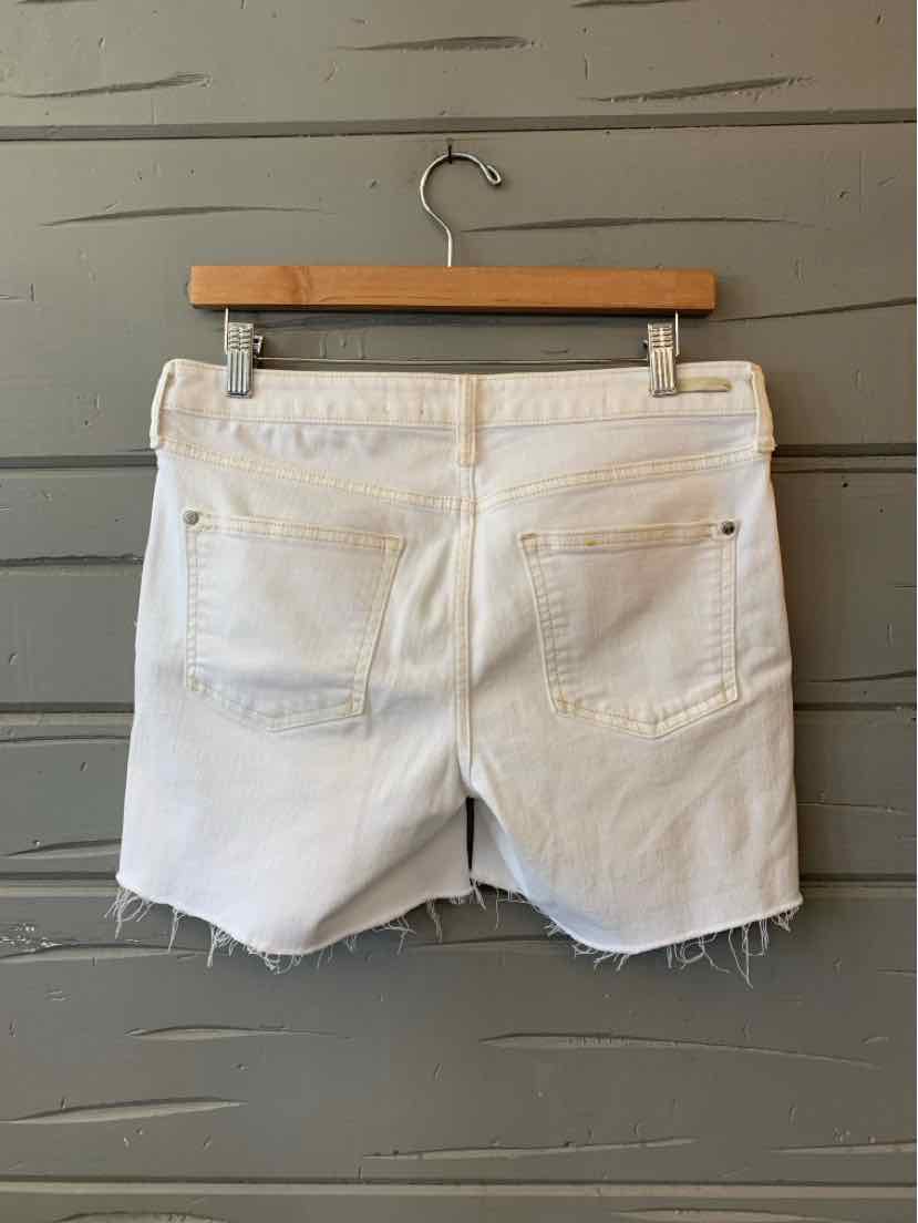 White denim shorts with raw hem and back pockets hanging on wooden hanger against gray wall.
