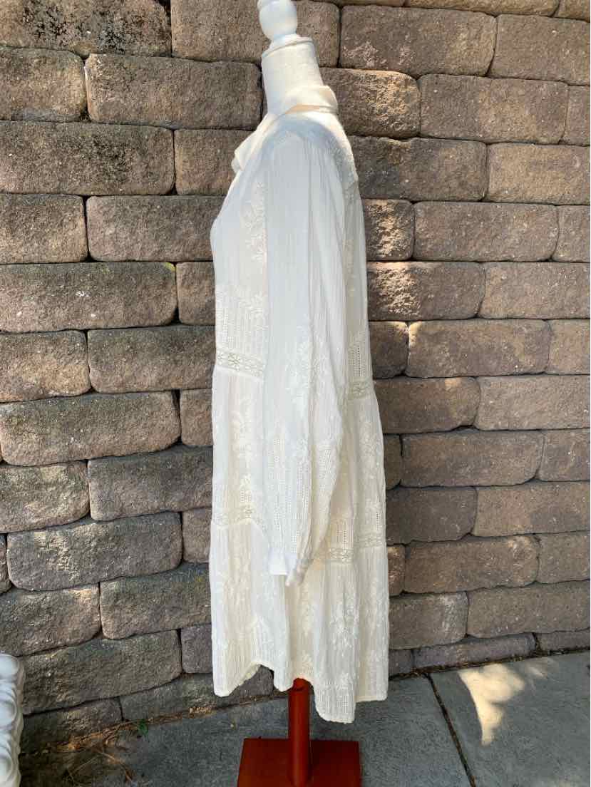 Long white cotton dress with long sleeves and loose, flowing fit on mannequin against stone wall background.