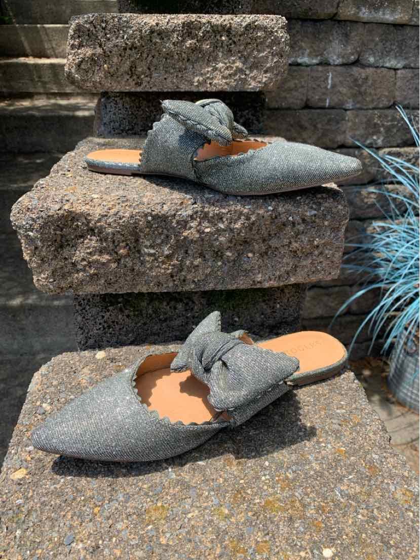Gray pointed-toe slip-on flats with knotted fabric detail and scalloped edges on stone steps outdoors.