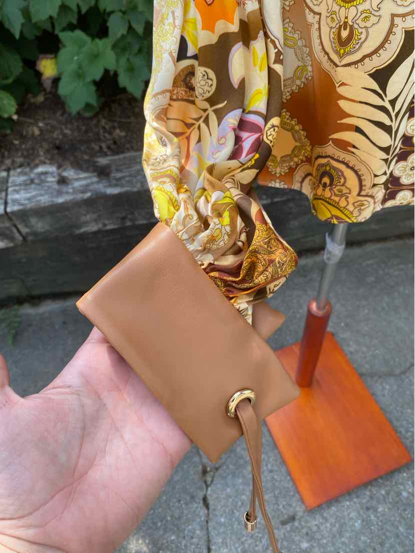 Tan leather wristlet with gold grommet and adjustable leather strap, held in hand outdoors.