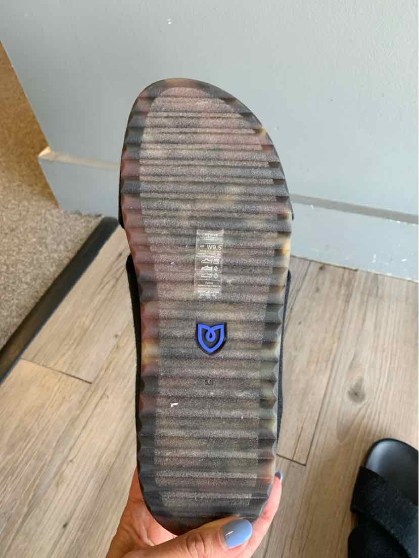 Black flat sole sandal with textured grip and small blue shield logo on bottom.