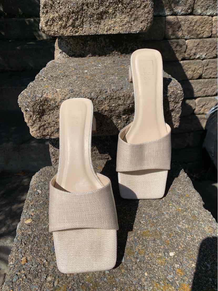Pair of beige slip-on sandals with wide fabric straps and square toes placed on stone steps.