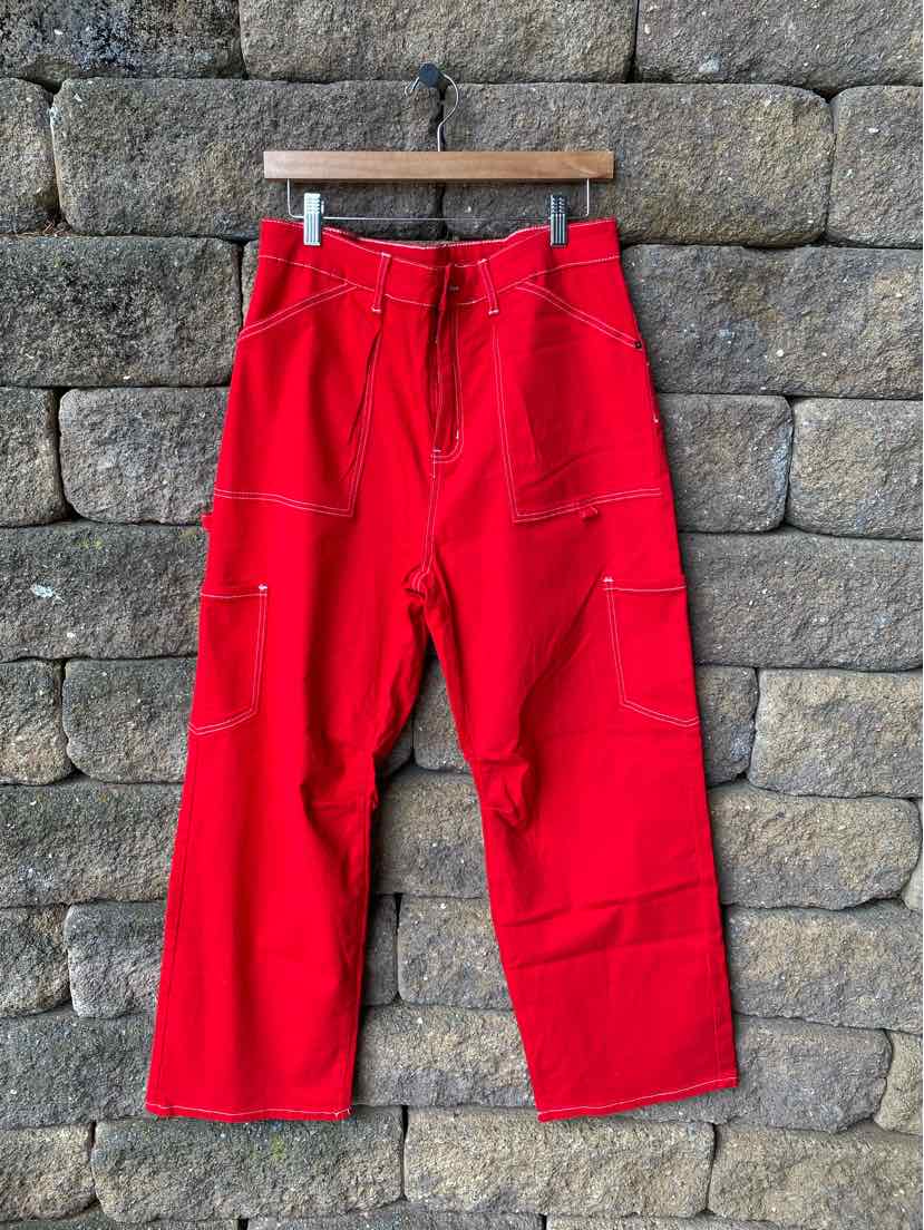 Red wide-leg pants with contrast white stitching and multiple pockets hanging on wooden hanger against stone wall.