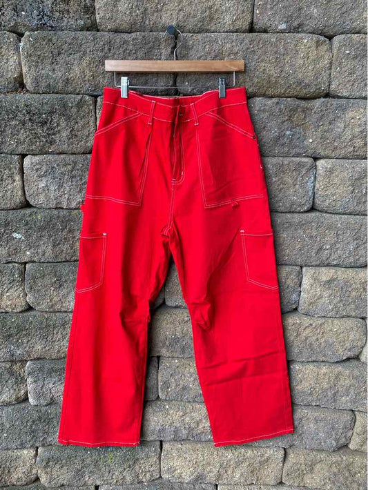 Red wide-leg pants with contrast white stitching and multiple pockets hanging on wooden hanger against stone wall.
