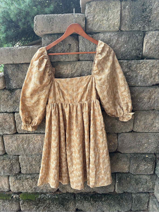 Brown jacquard women's dress with puff sleeves and square neckline hanging on wooden hanger against stone wall.