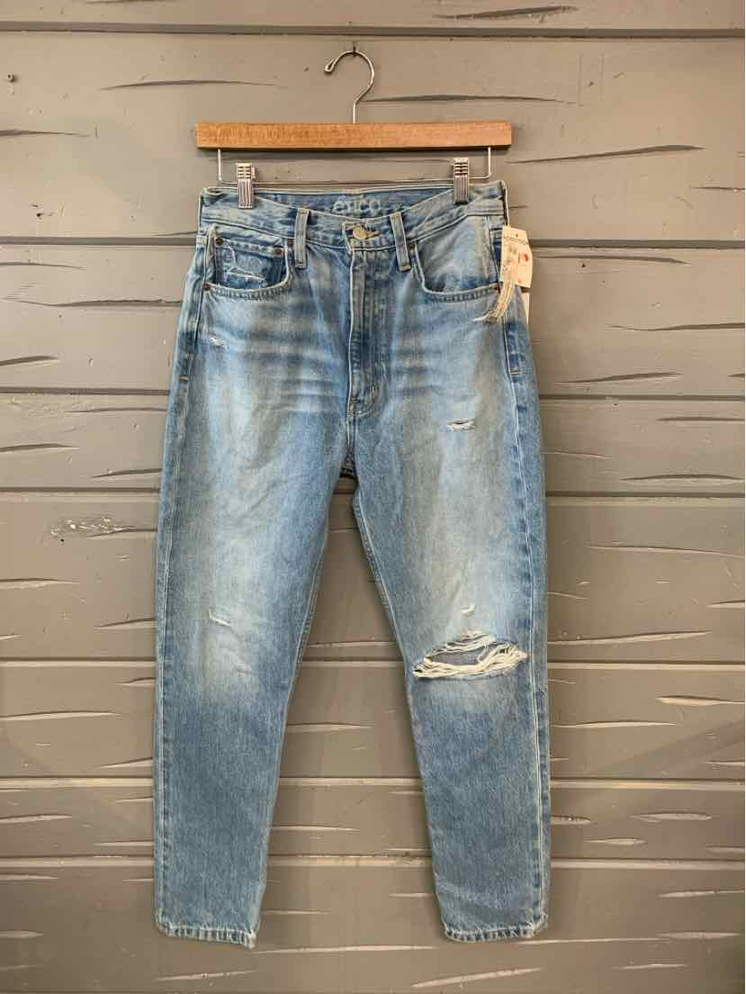 Light blue denim jeans with distressed knee and classic five-pocket design hanging on wooden hanger against gray wooden wall.