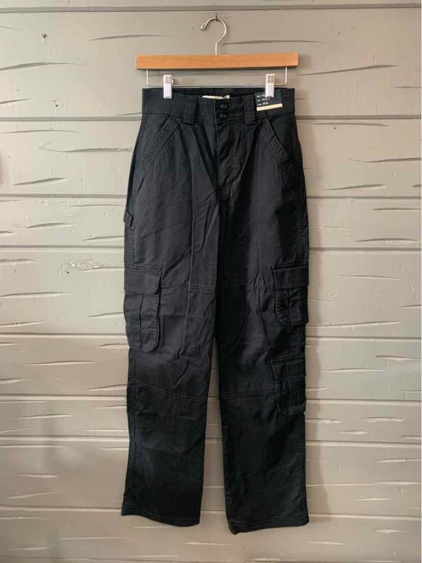 Black straight-leg pants with belt loops and side cargo pockets hanging on wooden hanger against light wood panel background.
