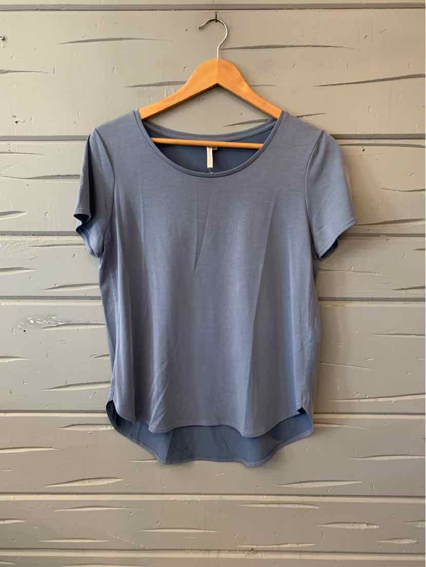 Light blue short-sleeve scoop neck blouse with curved hem on wooden hanger against gray wood background.