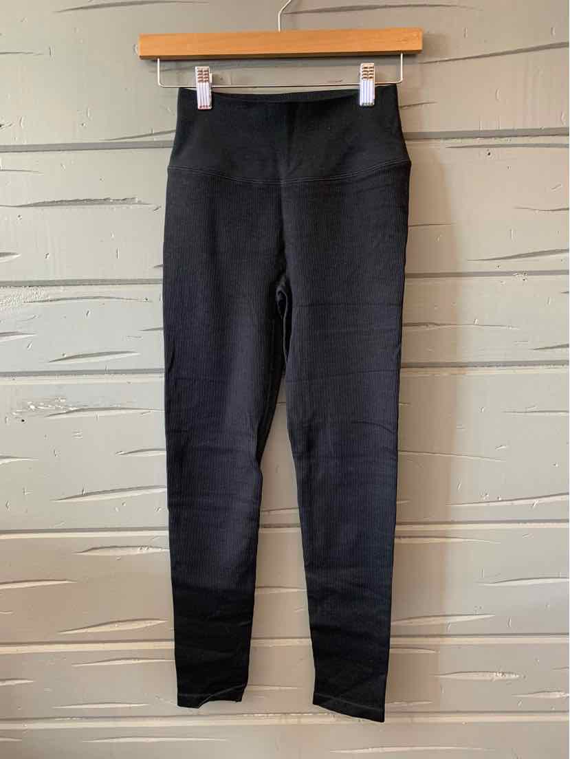 Black slim-fit knit leggings with a high waistband, size XS, hanging on wooden hanger against textured wall.