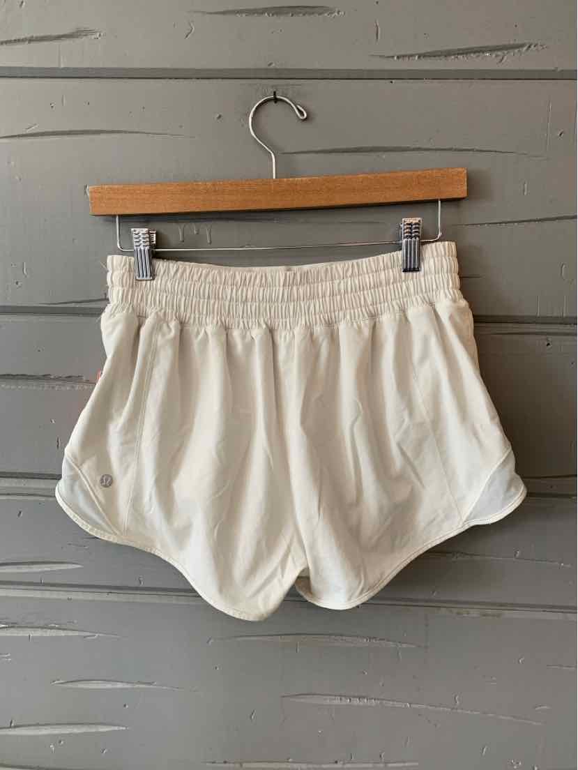 Light beige athletic shorts with elastic waistband and curved hem on wooden hanger against gray wall.