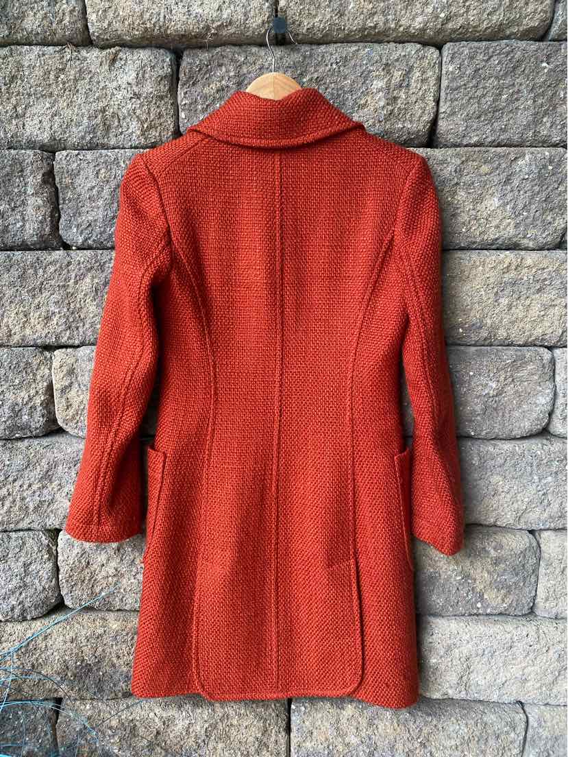 Orange wool blend long coat with tailored seams and notch collar hanging on wooden hanger against stone wall.