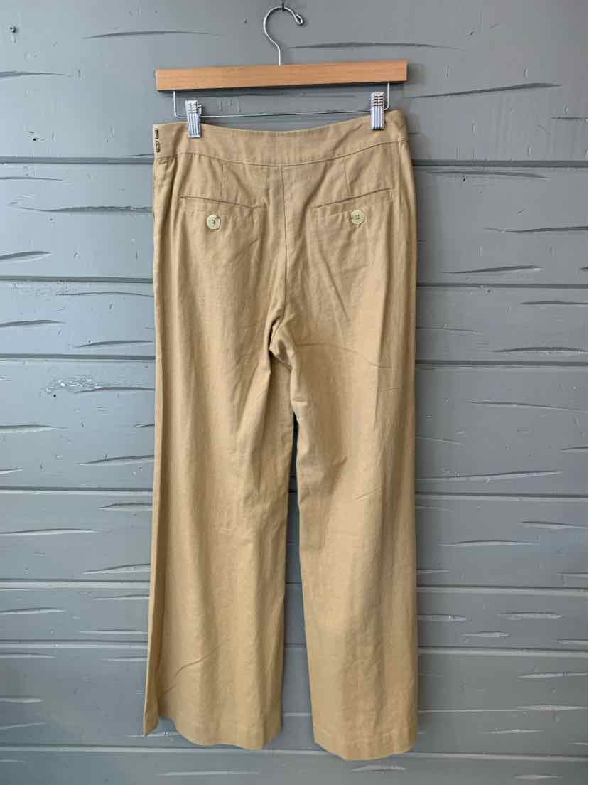 Tan high-waisted straight-leg pants with back buttoned pockets hanging on wooden hanger against gray wooden background.