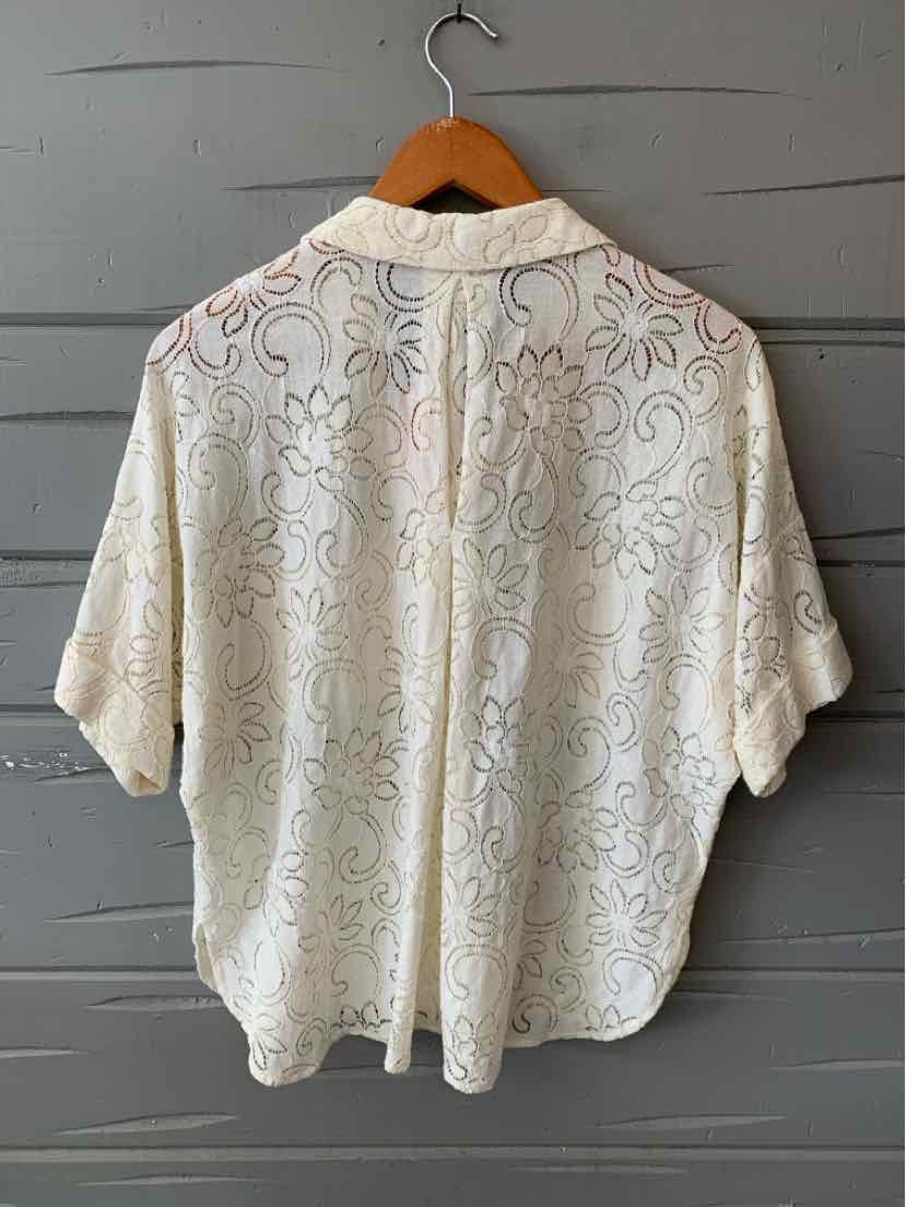 Cream-colored short-sleeve blouse with delicate floral lace pattern and relaxed fit hanging on wooden hanger.