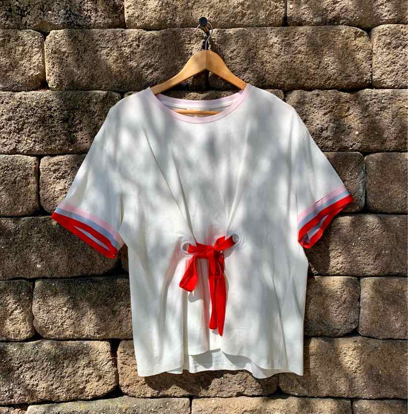 White short-sleeve T-shirt with red trim on sleeves and red tie detail at waist, hanging on wooden hanger against stone wall.