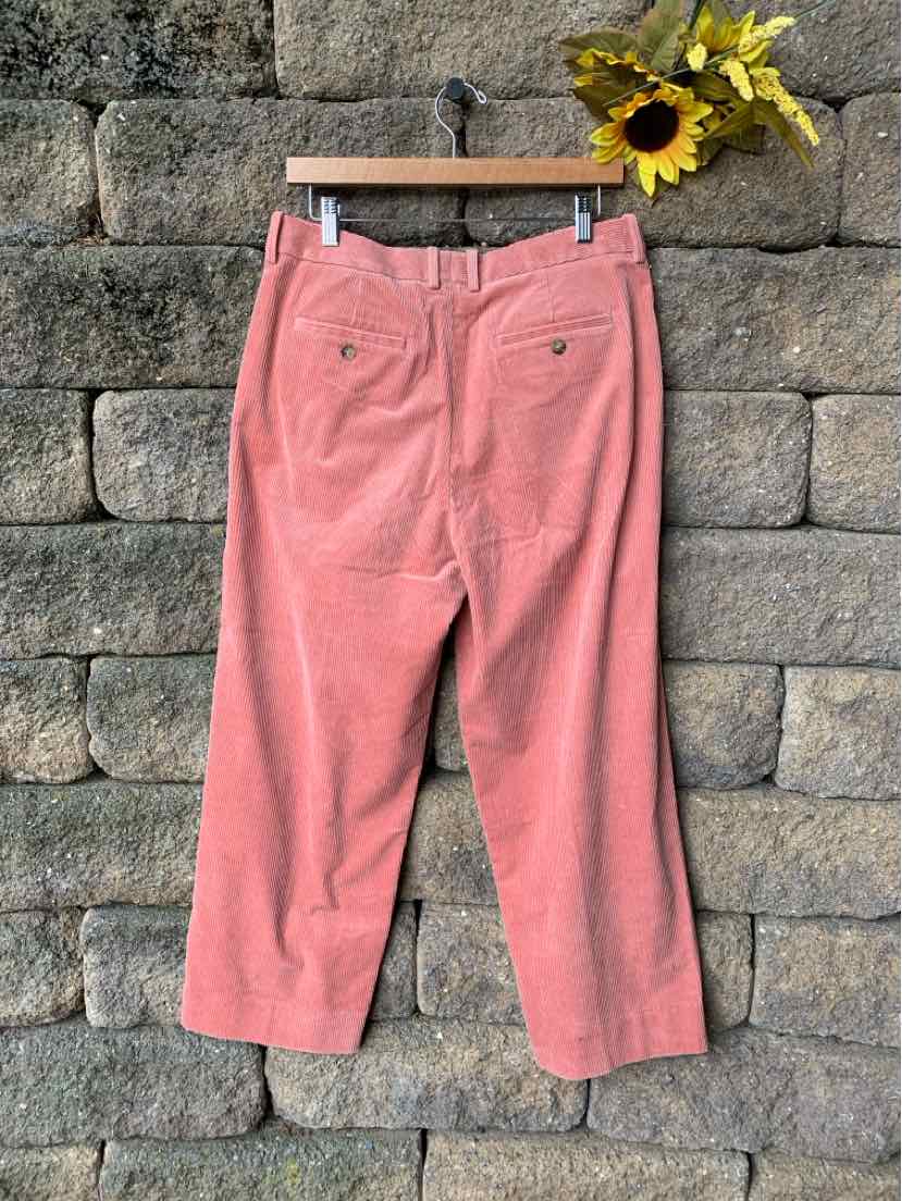 Pink corduroy pants with belt loops and back welt pockets hanging on a wooden hanger against a stone wall.