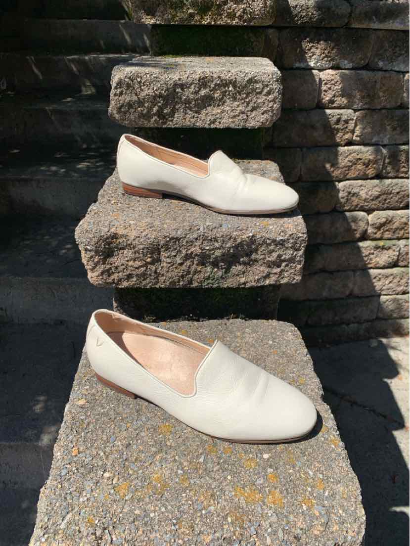 Cream-colored leather slip-on flats with rounded toes and low heels, displayed outdoors on stone steps.