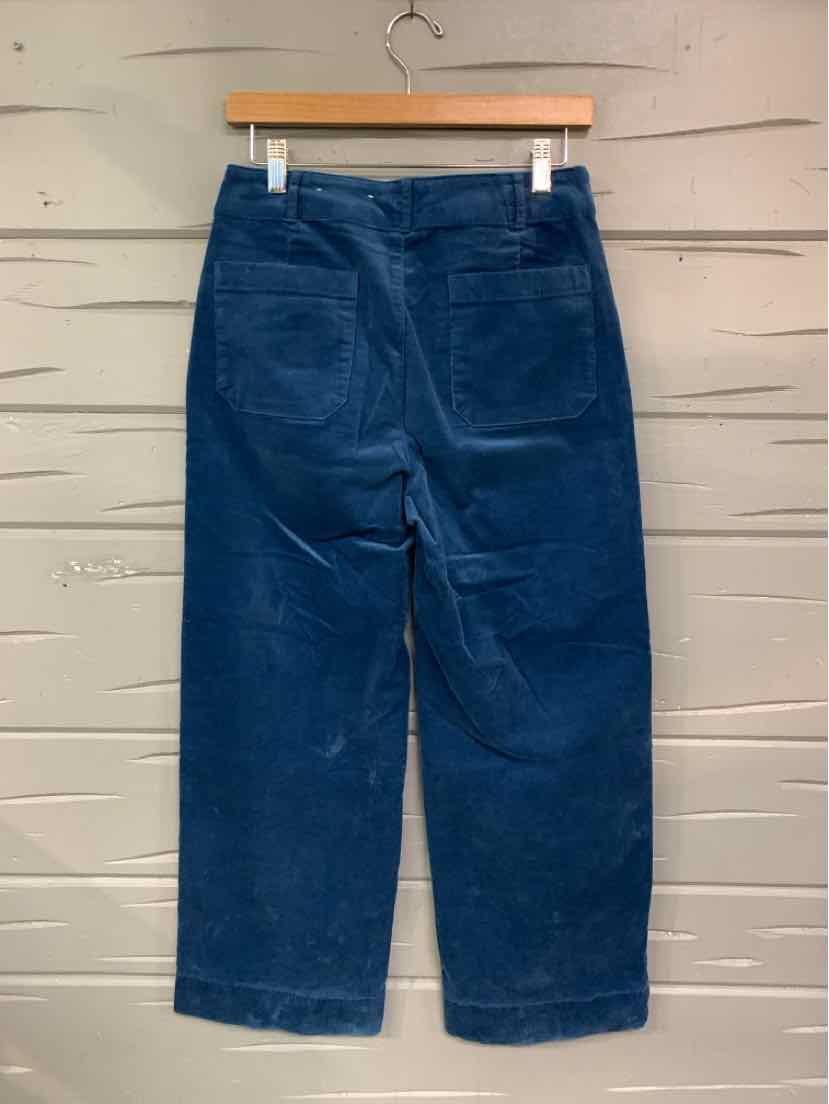 Wide-leg navy blue corduroy pants with back patch pockets hanging on wooden hanger against white wooden wall.